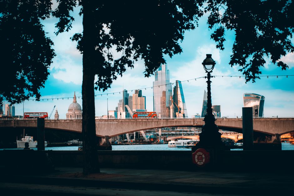 A photograph of a river scene at sunset showing a large bridge with ornate lampposts spanning across the water. In the background, a modern, curved glass skyscraper rises behind the bridge, reflecting
