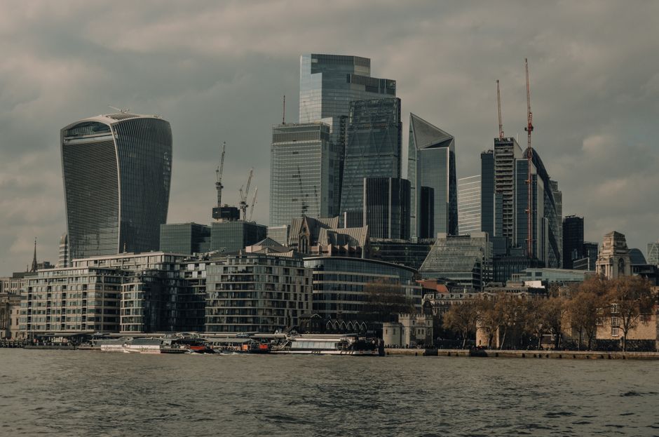 A panoramic view of a modern city skyline along a river, featuring high-rise office buildings and skyscrapers with glass facades, some under construction with cranes visible atop. In the foreground, there are mid-rise residential apartments with balconies, situated directly along the riverbank, which has boats and watercraft moored or navigating nearby. The scene is captured during the daytime under a cloudy sky, with natural light reflecting off the buildings. This image illustrates urban development and the transport of household items associated with residential relocations, aligning with home moving services offered by Waterloo Removals, particularly when coordinating furniture transport and packing during a relocation process in SE1 areas like Lower Marsh.